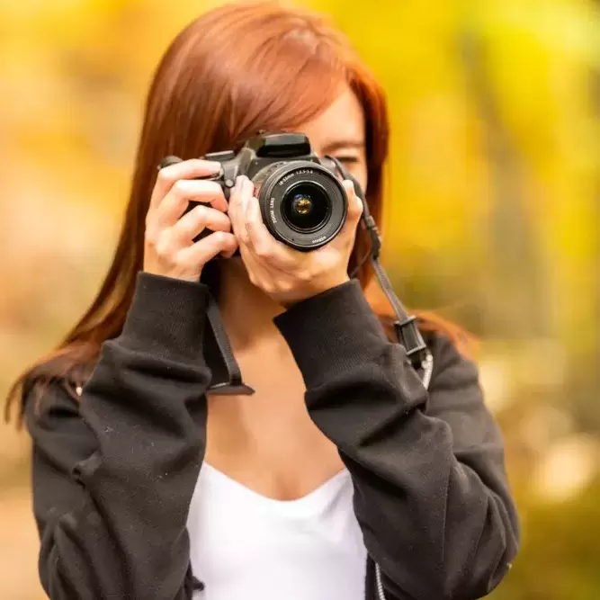 A young woman with reddish hair takes pictures with a reflex camera in an autumnal forest of yellow and other colors