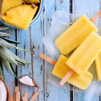 Pina colada popsicles on a table with ice, half a coconut, and a bowl of sliced pineapple. 