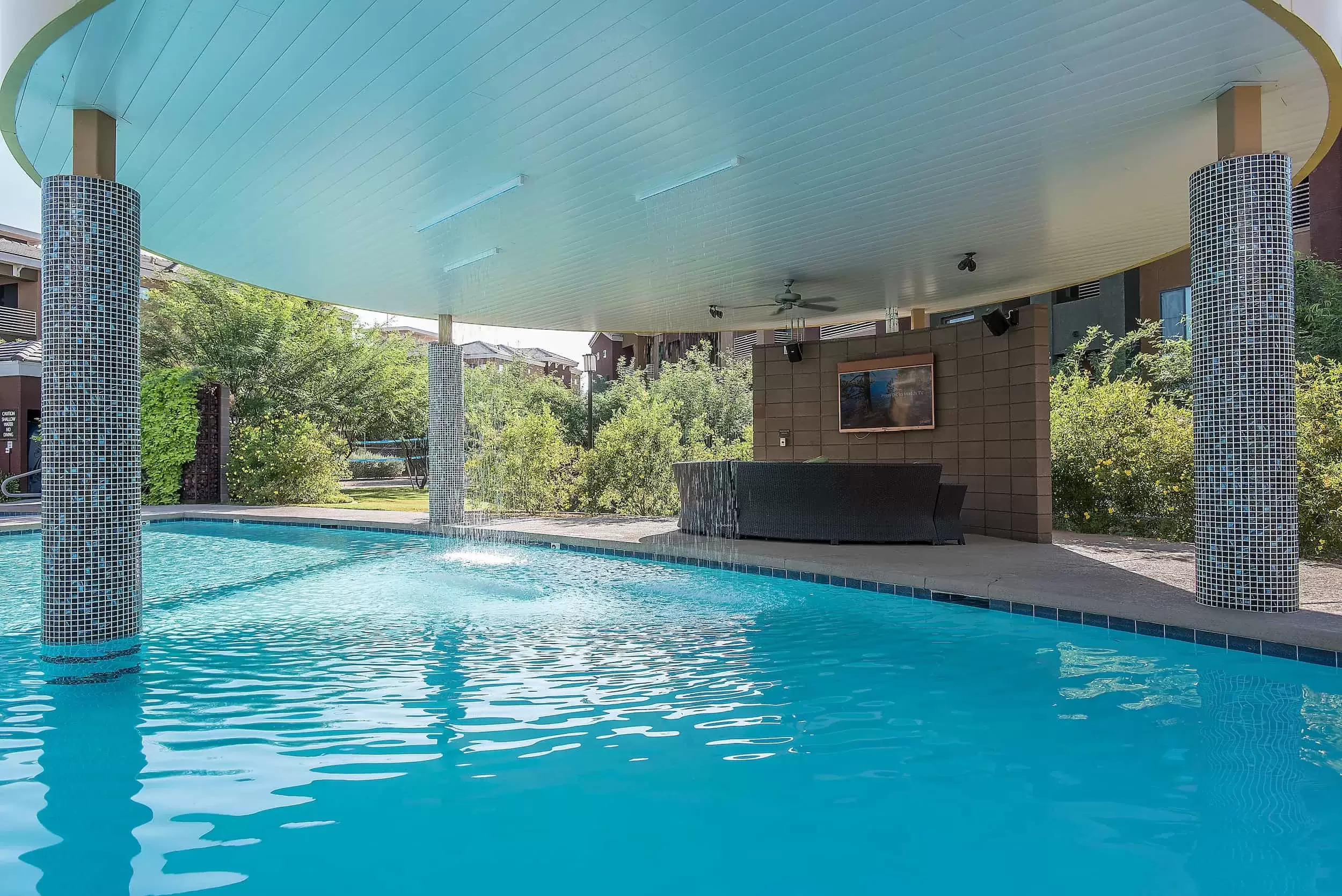 Water falls from overhead into the pool, beside that a couch sets in the shade in front of a large screen tv.